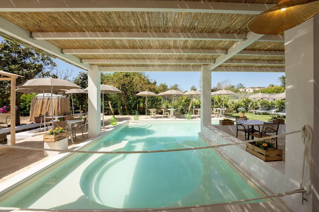 A luxurious covered poolside area with turquoise water, white pergola with woven ceiling details, lounge umbrellas, and manicured garden views in the background.