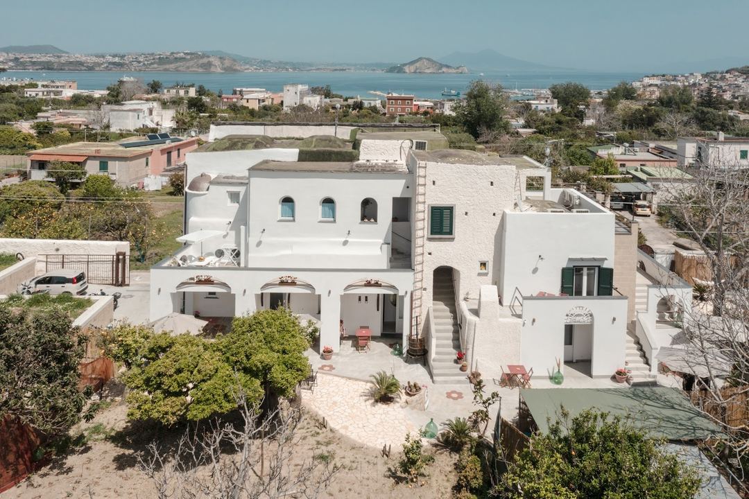Aerial view of a whitewashed Mediterranean-style property with terraced courtyards overlooking a coastal bay and distant mountains.