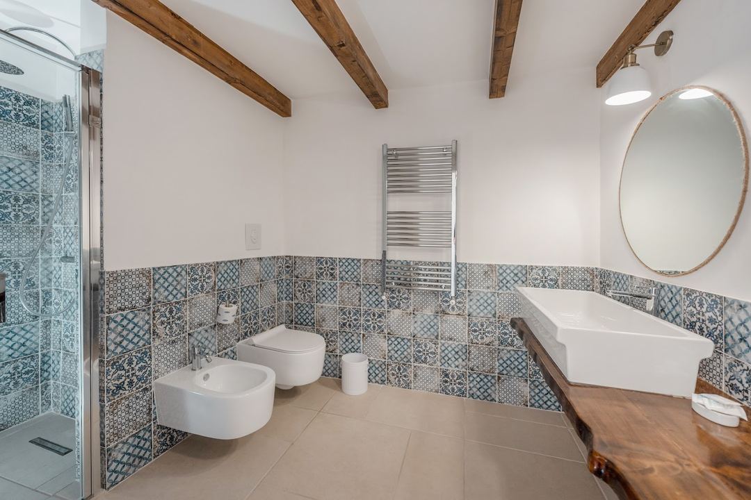 A spacious bathroom featuring decorative blue patterned tile wainscoting, dual wall-mounted fixtures, a wooden vanity with vessel sink, and exposed wooden ceiling beams.