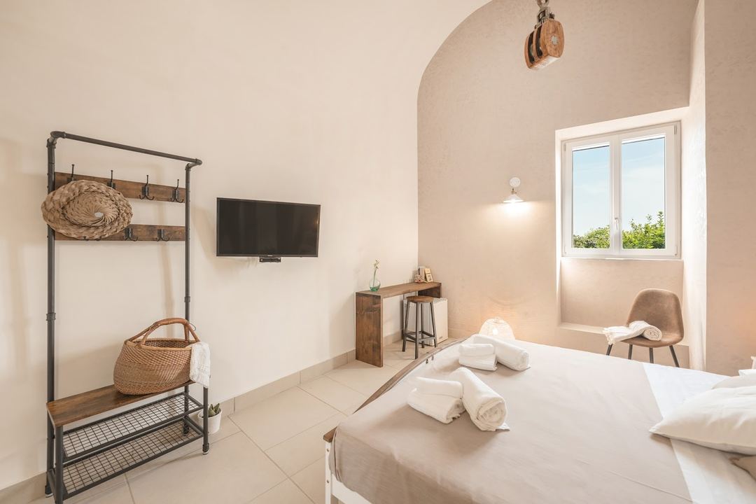 A bright, minimalist bedroom featuring a bed with rolled towels, a metal and wood clothing rack, wall-mounted TV, and a window with countryside views.