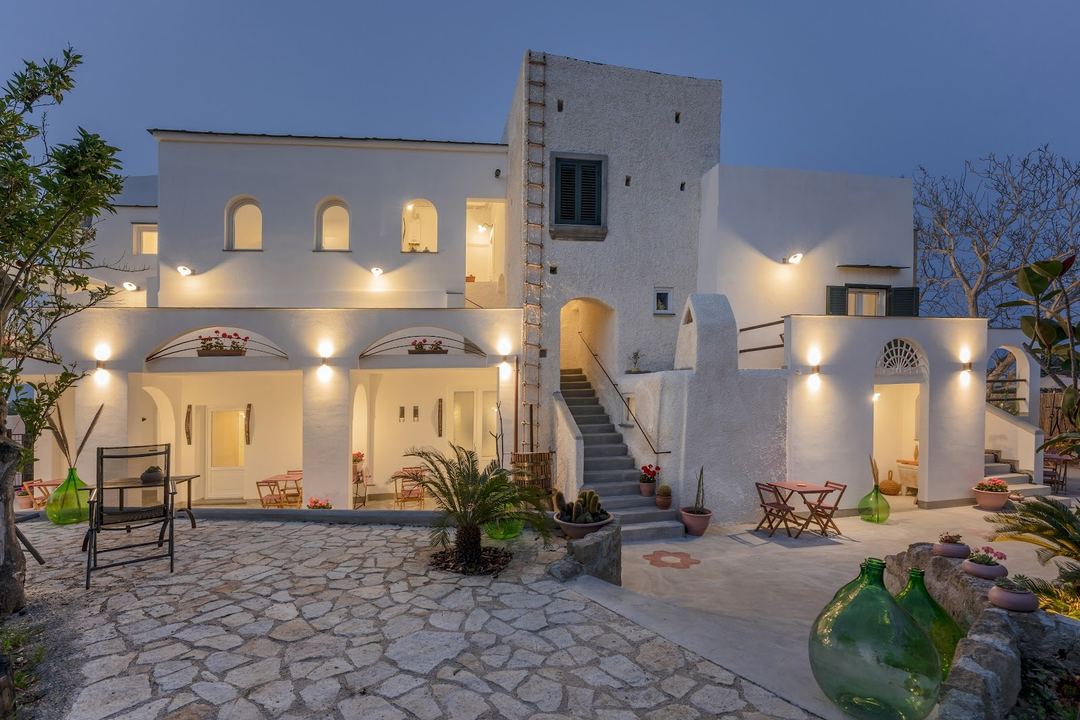 The whitewashed exterior of Calacala Rooms & Farm Experience is beautifully illuminated at dusk, showcasing the traditional Mediterranean architecture with stone courtyard, potted plants, and welcoming seating areas.