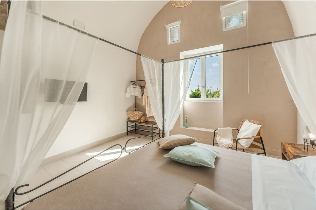 A bright, minimalist bedroom with an arched ceiling, sheer canopy bed, and floor-to-ceiling windows overlooking natural landscape.