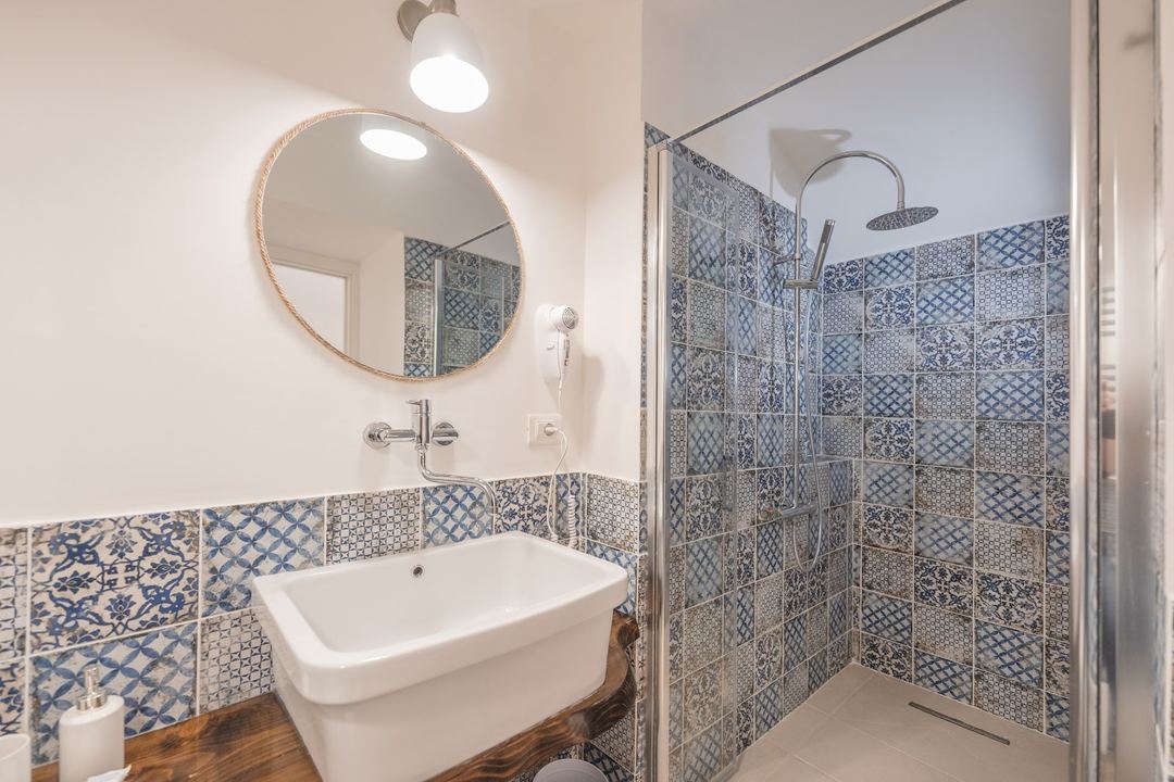 A modern bathroom featuring a white rectangular sink, round mirror with natural wood frame, and a walk-in shower with blue and white patterned tile work.