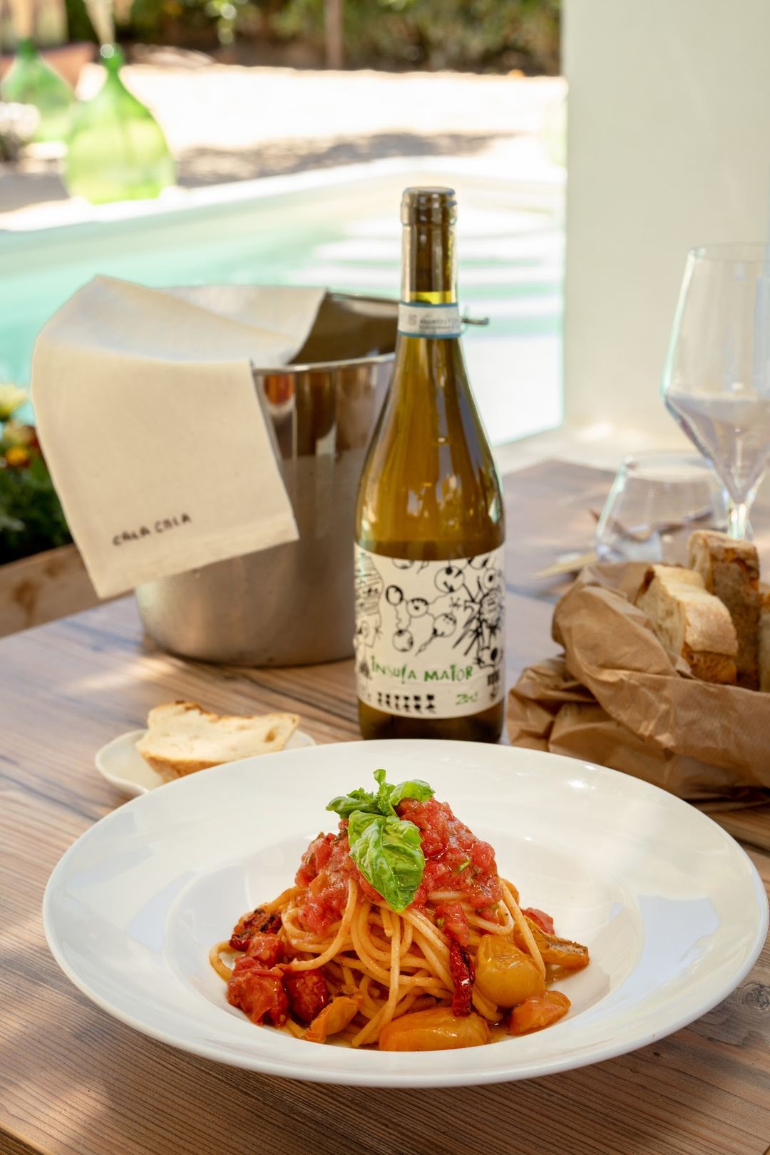 A plate of spaghetti with tomato sauce and fresh basil is served poolside with a white wine bottle and bread at Calacala Rooms & Farm Experience.