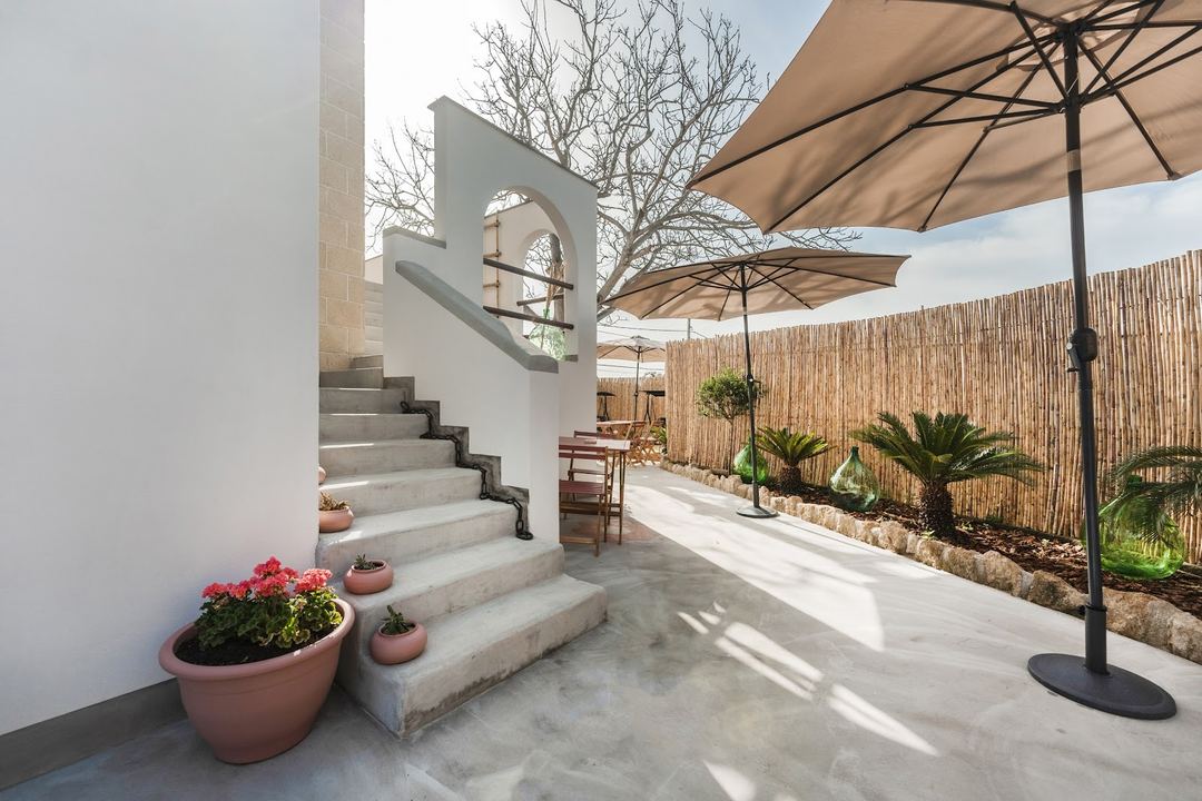 A sunlit courtyard patio featuring concrete stairs with architectural arch details, potted flowers, wooden umbrellas, and rustic bamboo fencing surrounding the outdoor seating area.