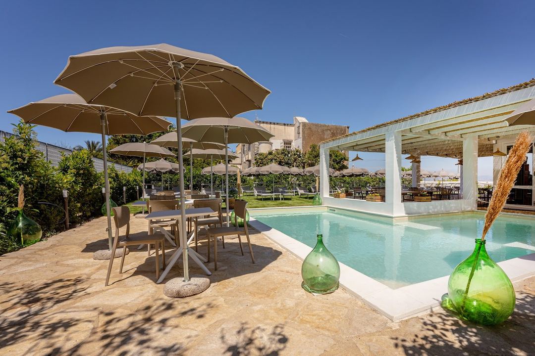 The hotel's swimming pool area features turquoise water, shaded umbrellas, dining tables, and white pergolas with views of the surrounding countryside.